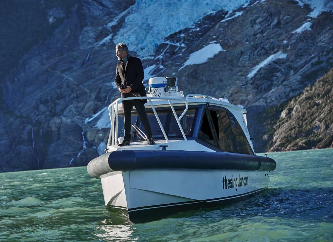 A man is standing at the tip of a fishing boat, surrounded by turquoise waters and snowy mountains. 