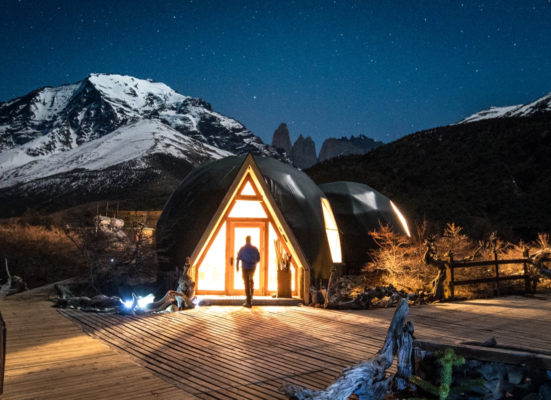 A man is walking into a dome at night, and the sky is illuminated with stars, and there is a snowy mountain in the background. 