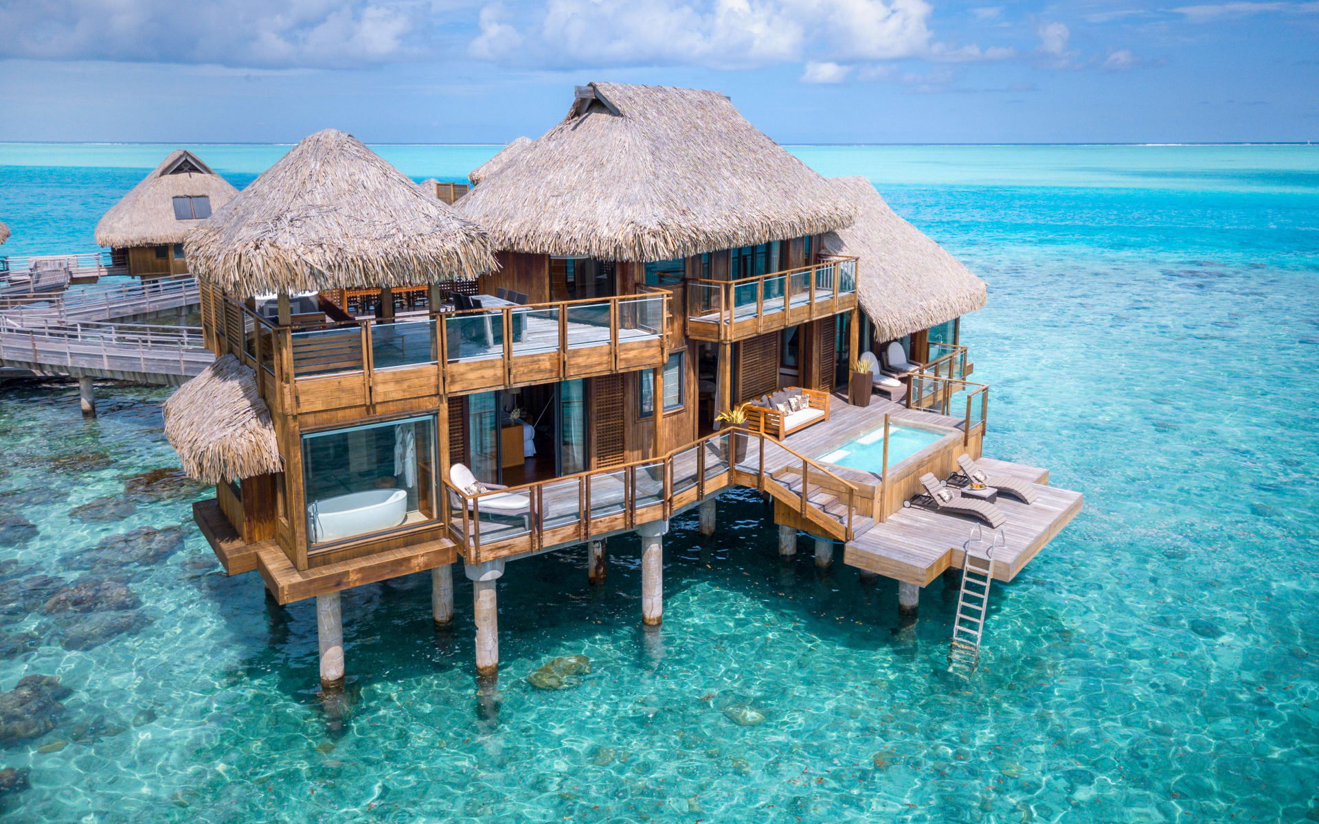 A thatched-roof villa has two floors with wraparound balconies, a pool and outdoor seating. 