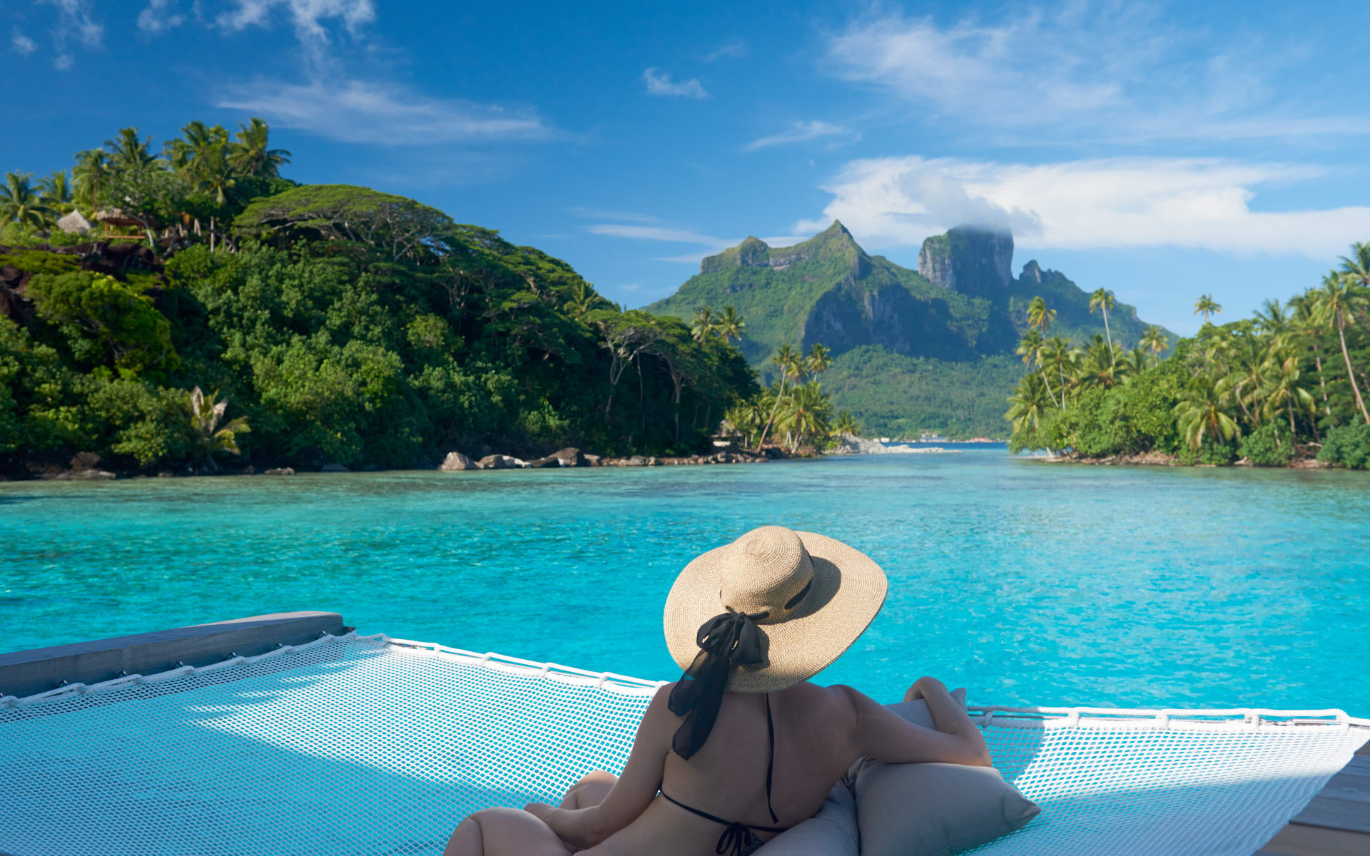 A woman in a straw hat is sitting on a net trampoline, looking out to the turquoise ocean and mountains. 