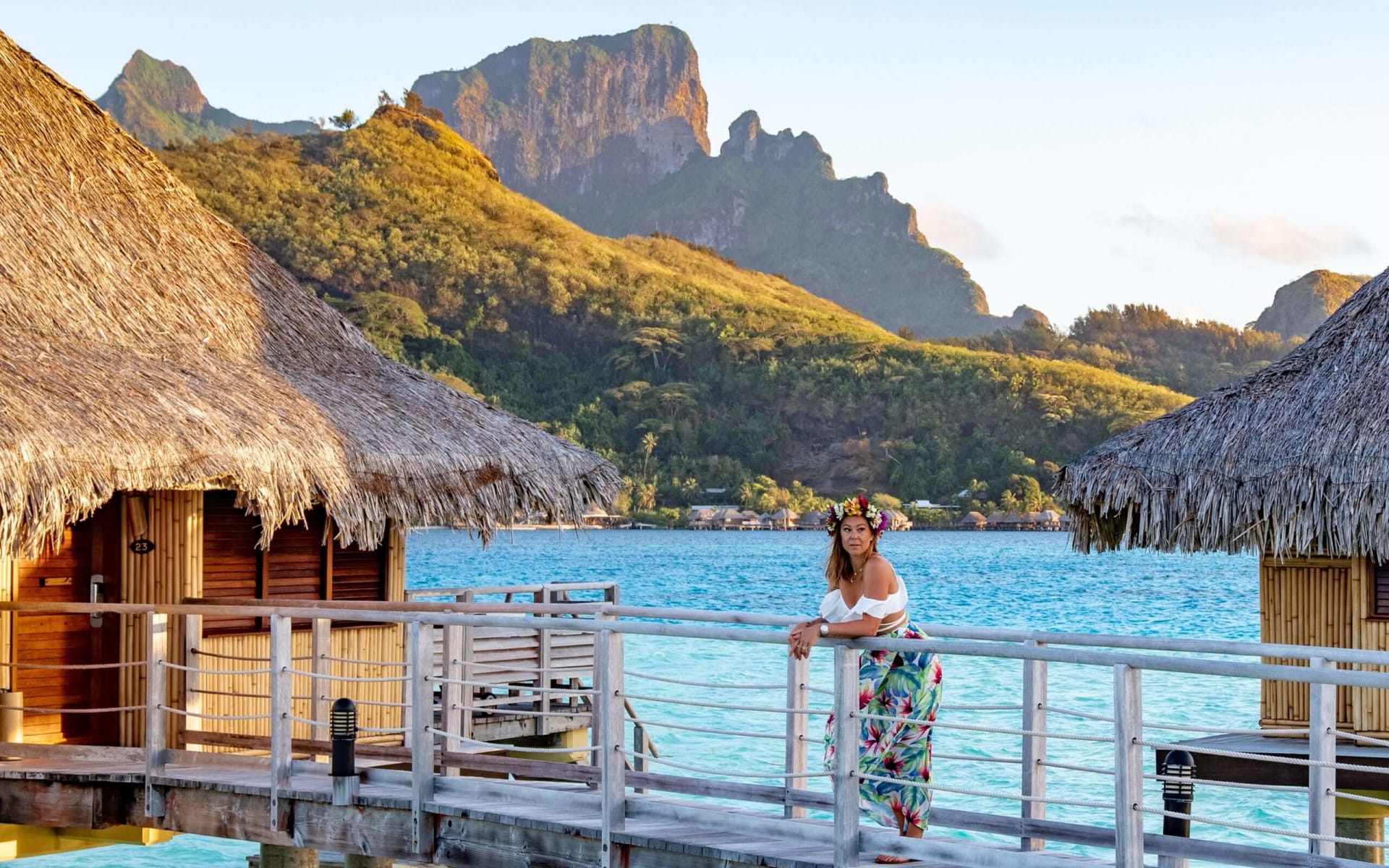 A woman is standing on a bridge backed by thatched-roof villas and a mountain. 