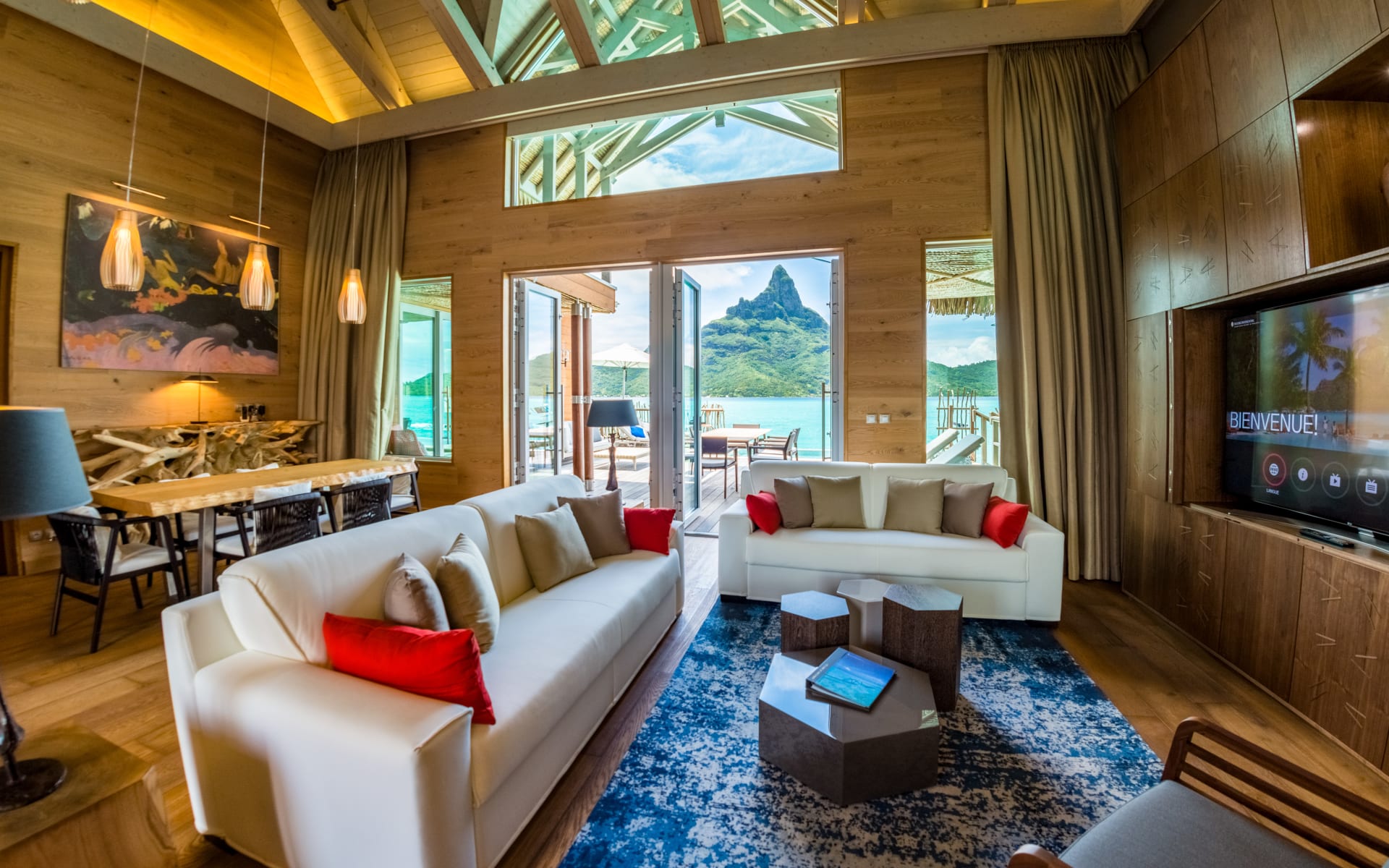 A large living room has two white sofas, a TV, a dining table and doors leading to a deck with a pool. 