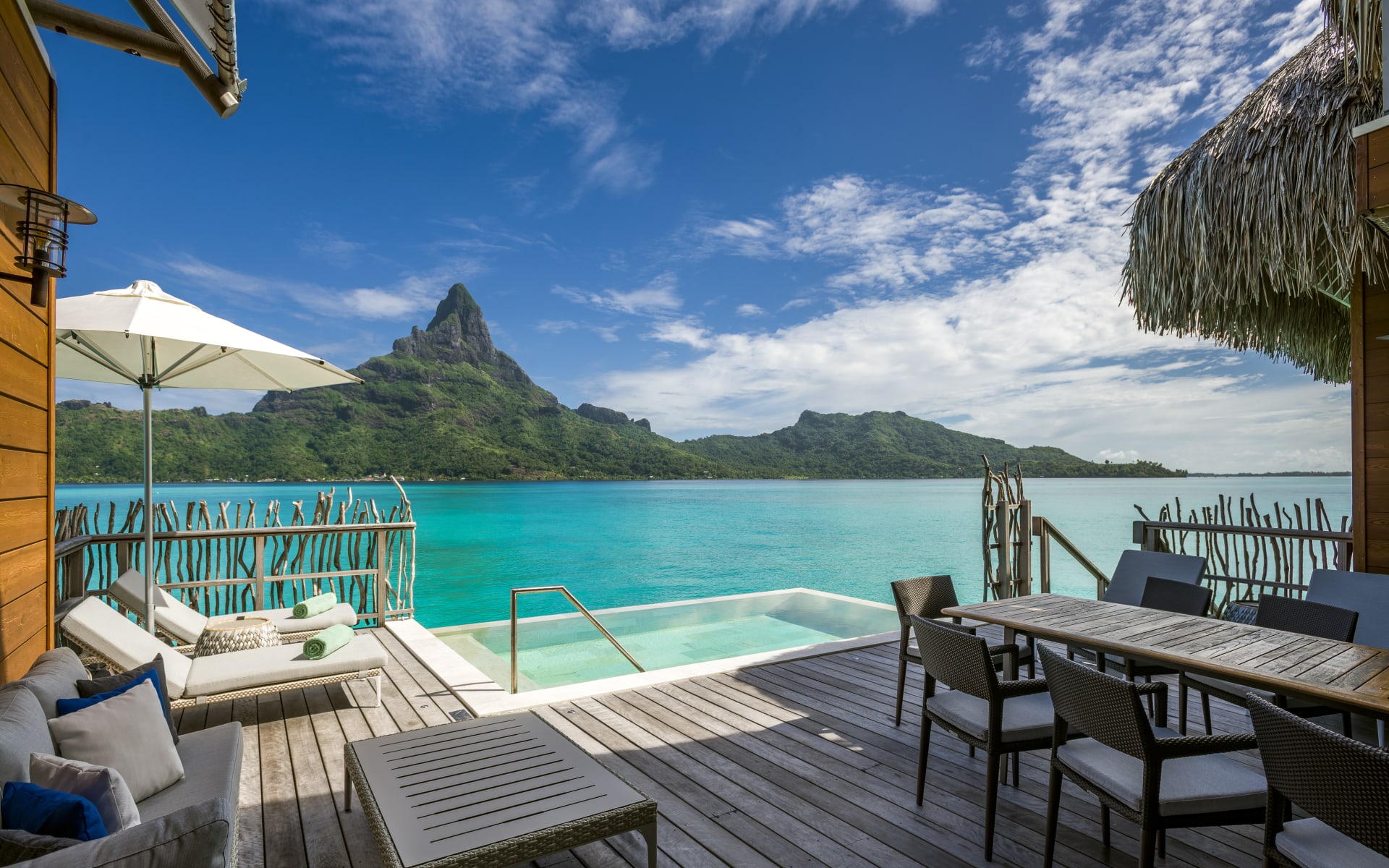 A wooden deck hanging over the ocean has sun loungers, a pool and a dining area. 