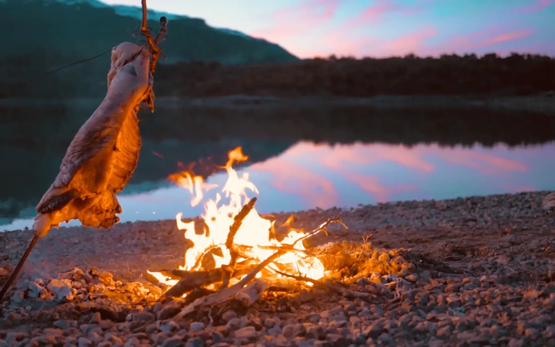 Some freshly-caught fish is being slow-cooked over a log fire, ahead of an evening scene of Lake San Martin.