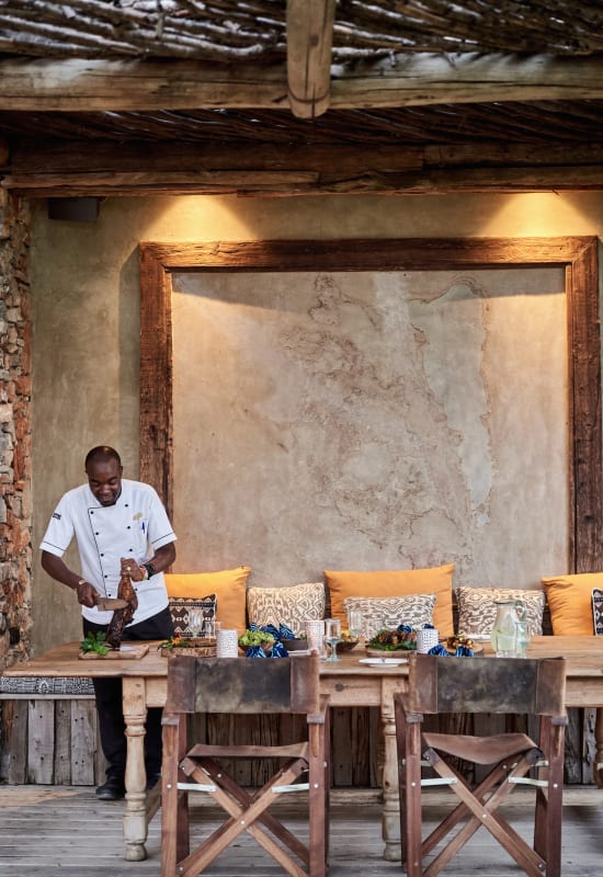 A friendly sever prepares a plentiful spread of barbecued meat on a wooden dining table surrounded by colourful cushions.