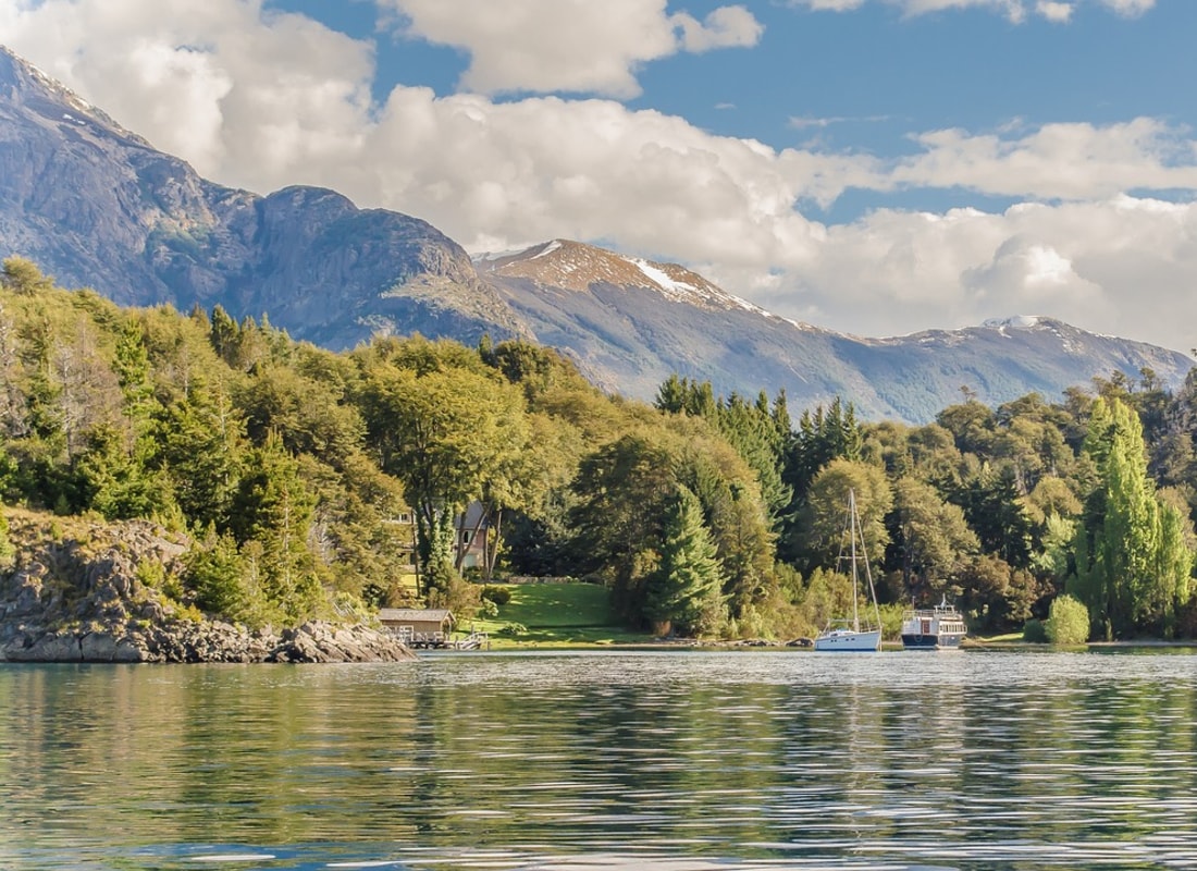 Sailing boats are docked on a gentle lake, surrounded by green trees and snow-capped mountains. 
