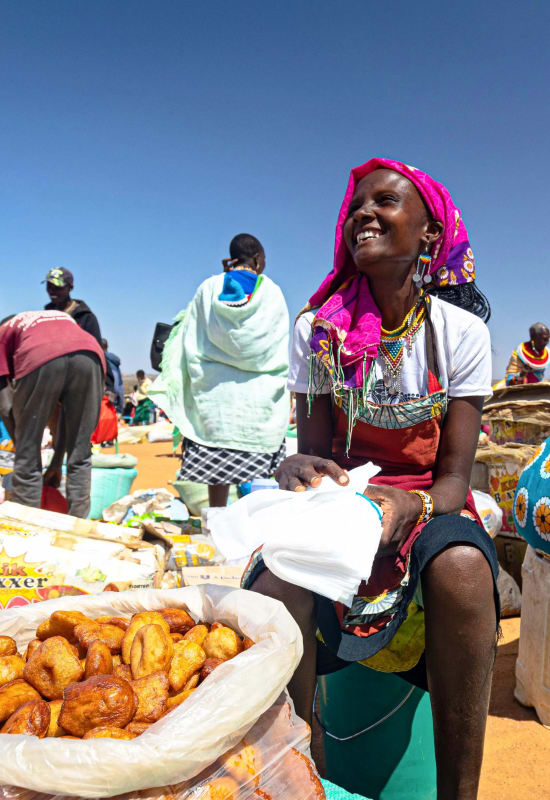 A local woman sits smiling beside a large container of freshly-cooked goods.