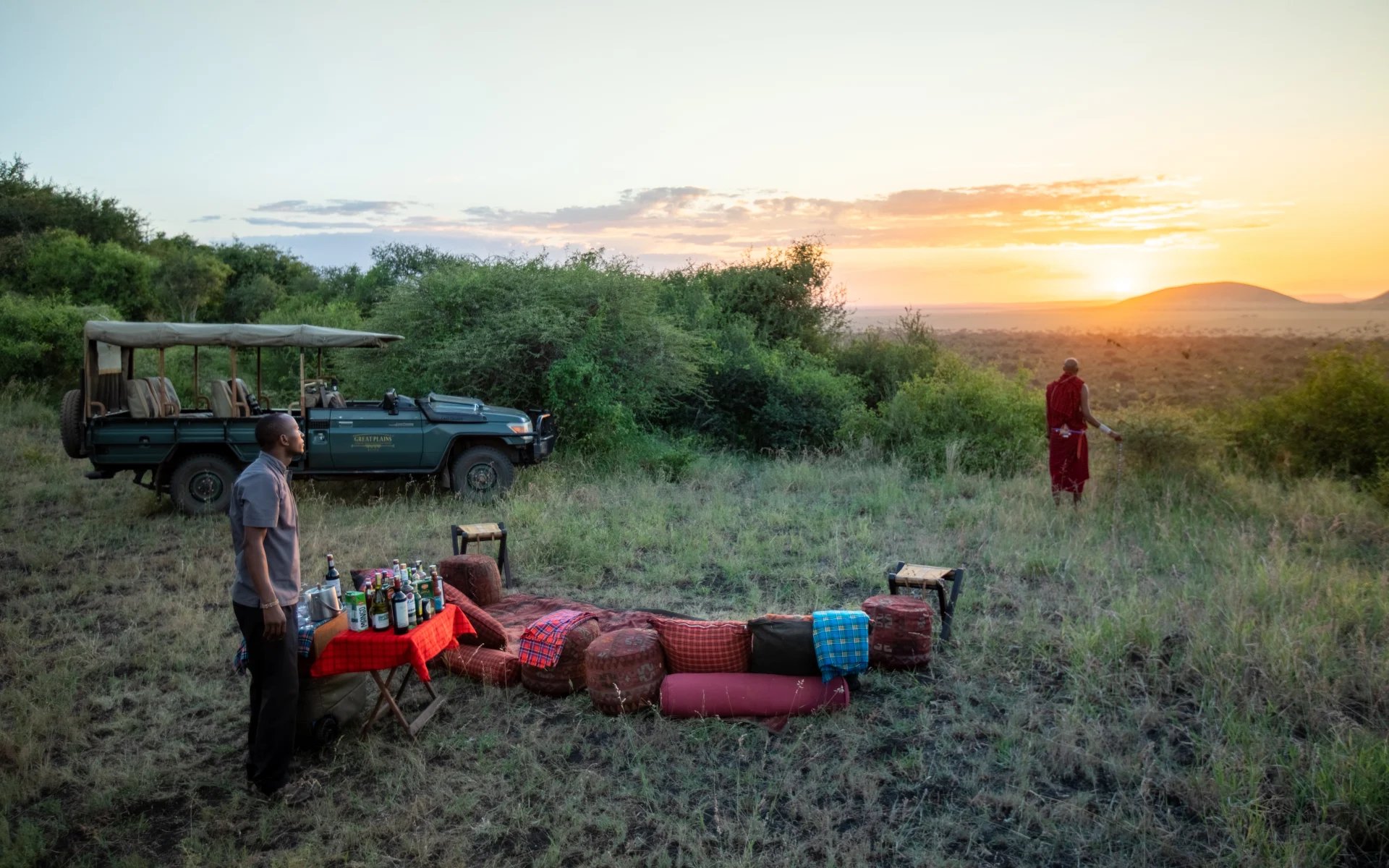 A picnic is set out on a hill on the reserve during sunset. A camp guide and local Maasai warrior admire the view