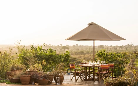 A wooden deck has a table and four chairs under a brown umbrella. It is all surrounded by bushes. 
