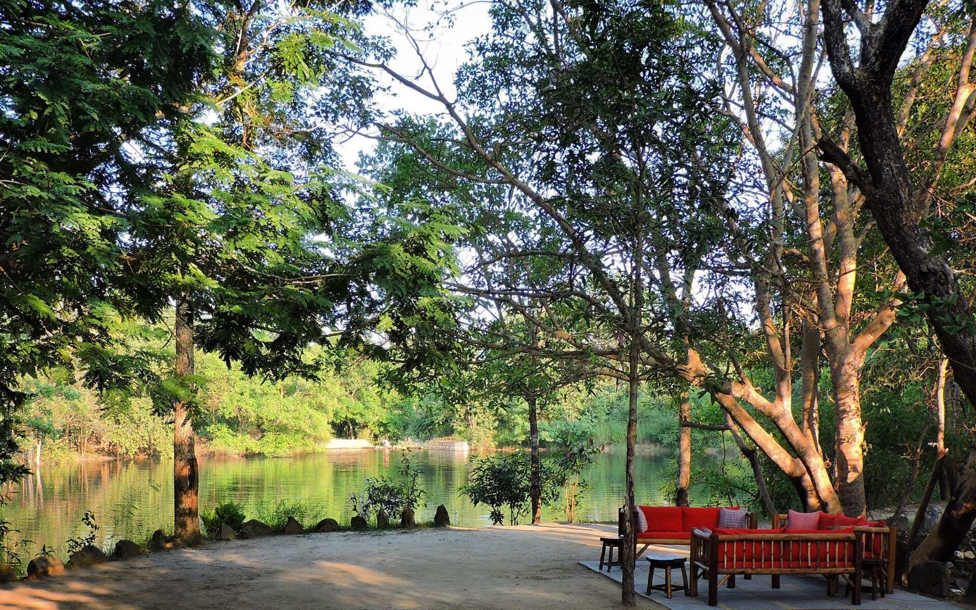 Red sofas face a body of water in Shergarh Tented Camp, surrounded by trees.