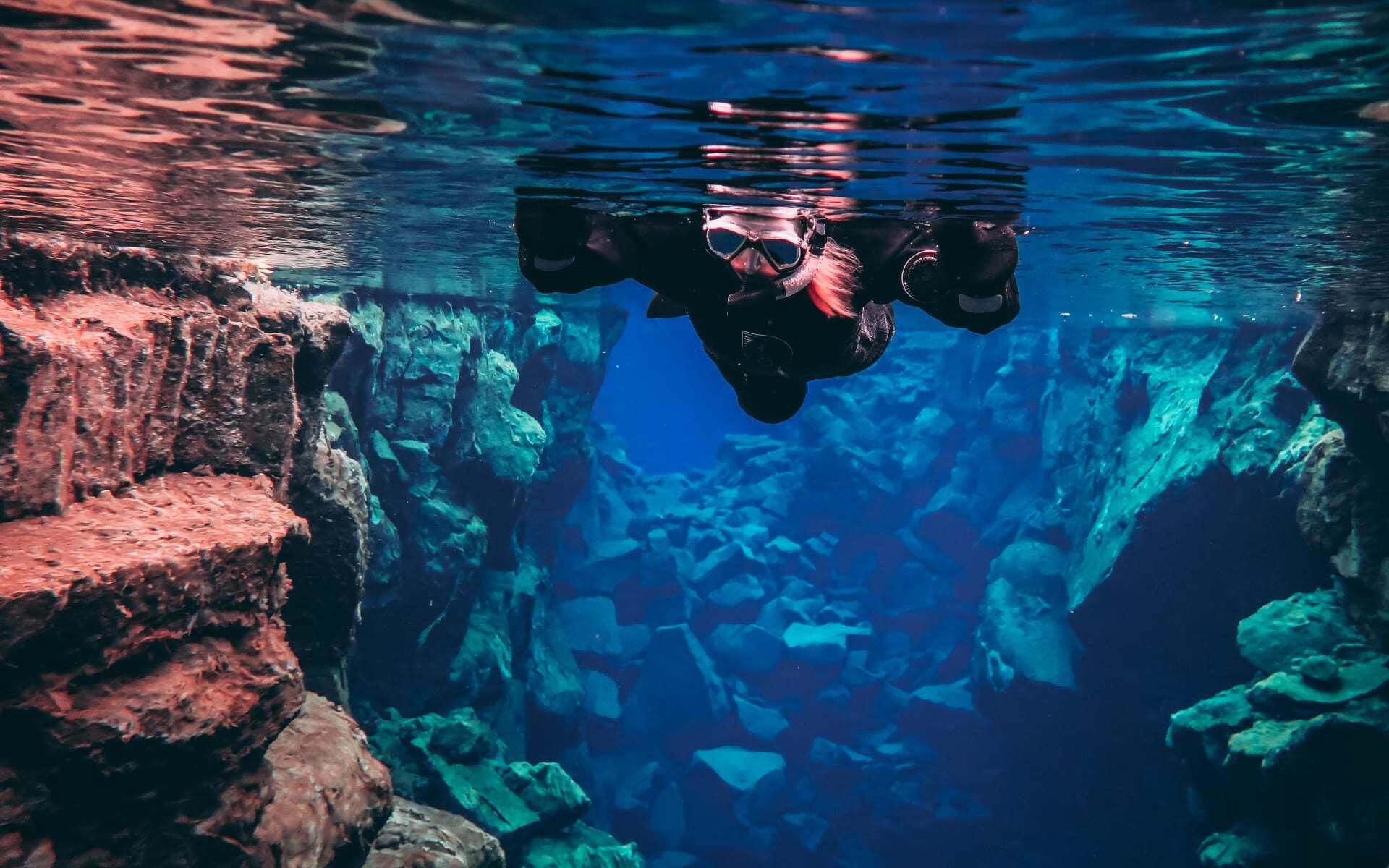 A lady snorkels in the Silfra Fissure, surrounded by rocks and deep blue water.