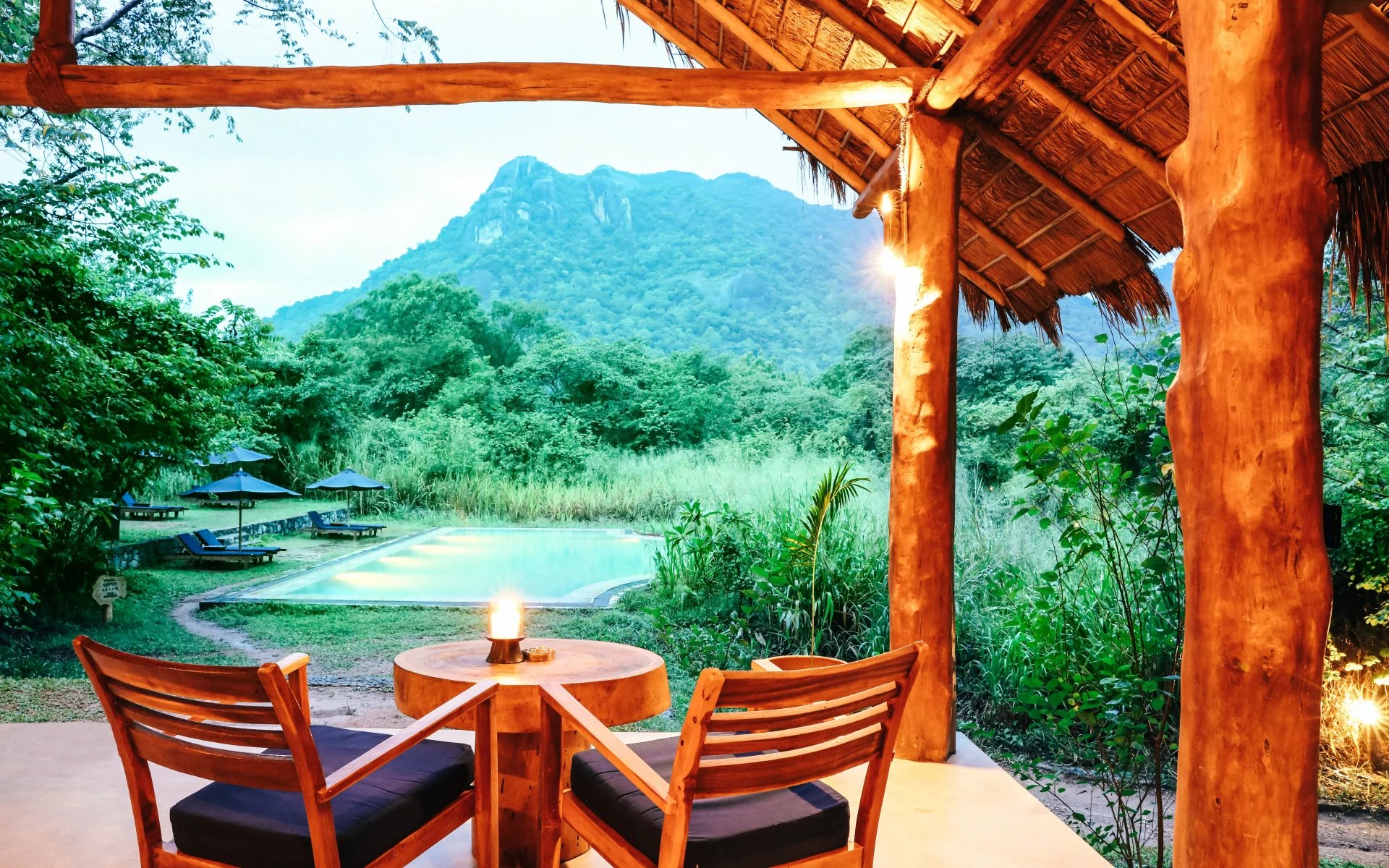 Two wooden chairs sit under a shelter, facing the lodge's outdoor pool and the mountains which rise behind it.