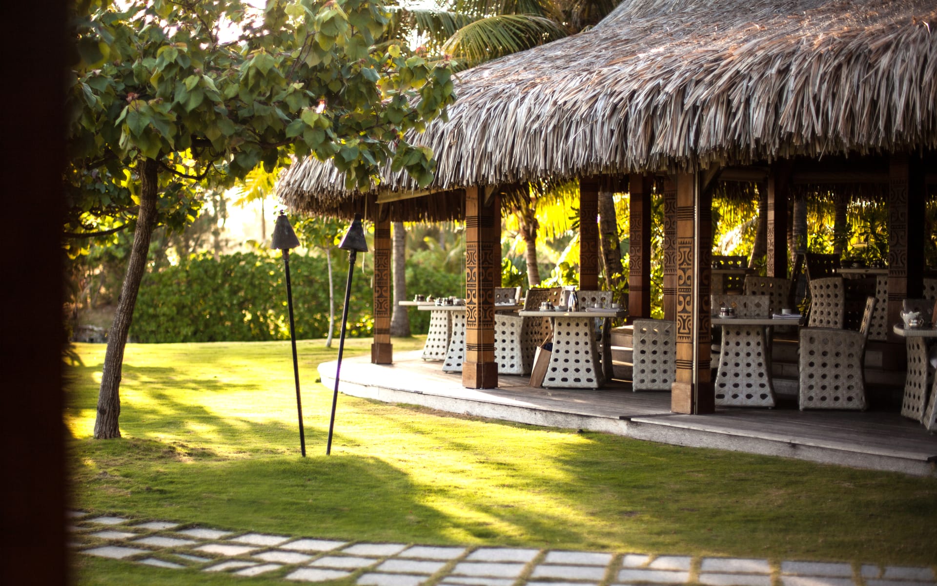 A thatched-roof building has tables and chairs overlooking a garden.
