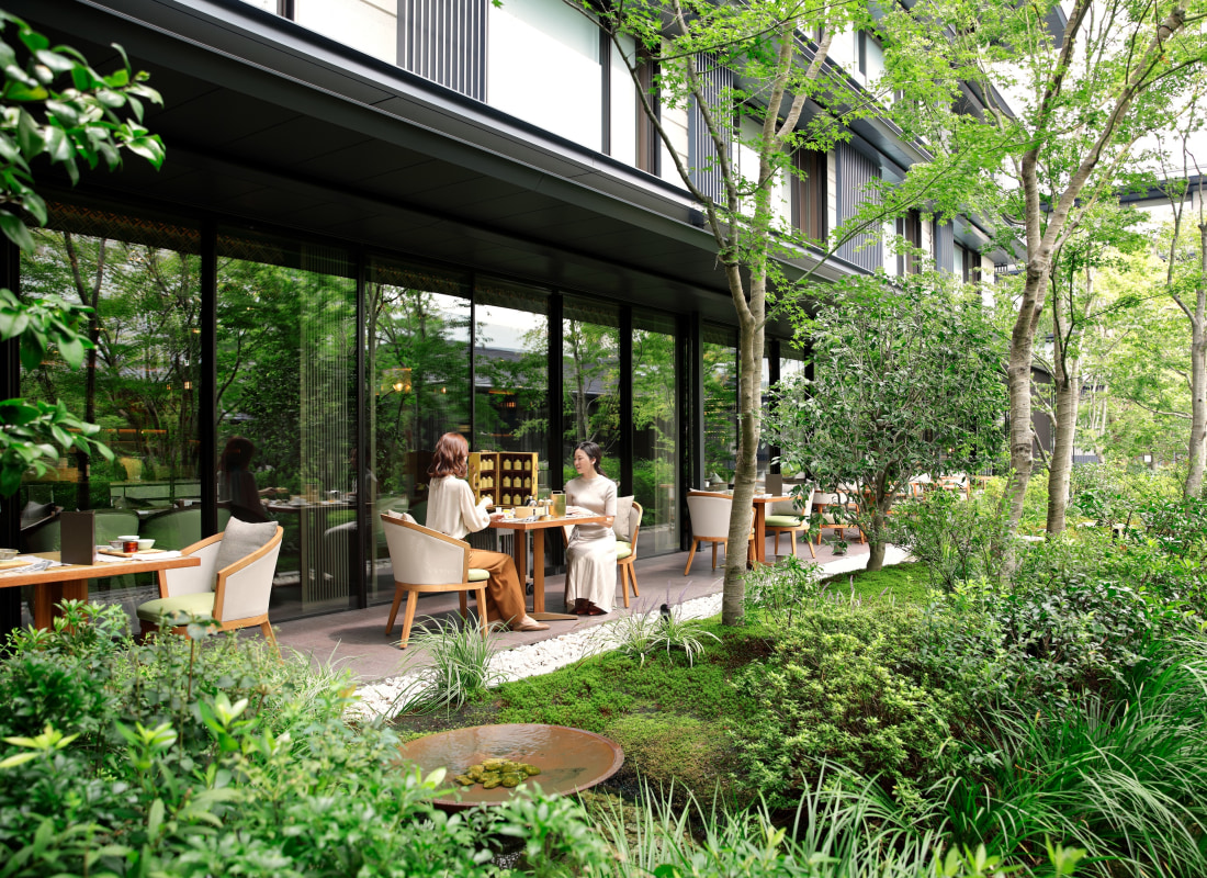 Two women are sitting at a wooden table on an outdoor balcony overlooking a green wilderness. 