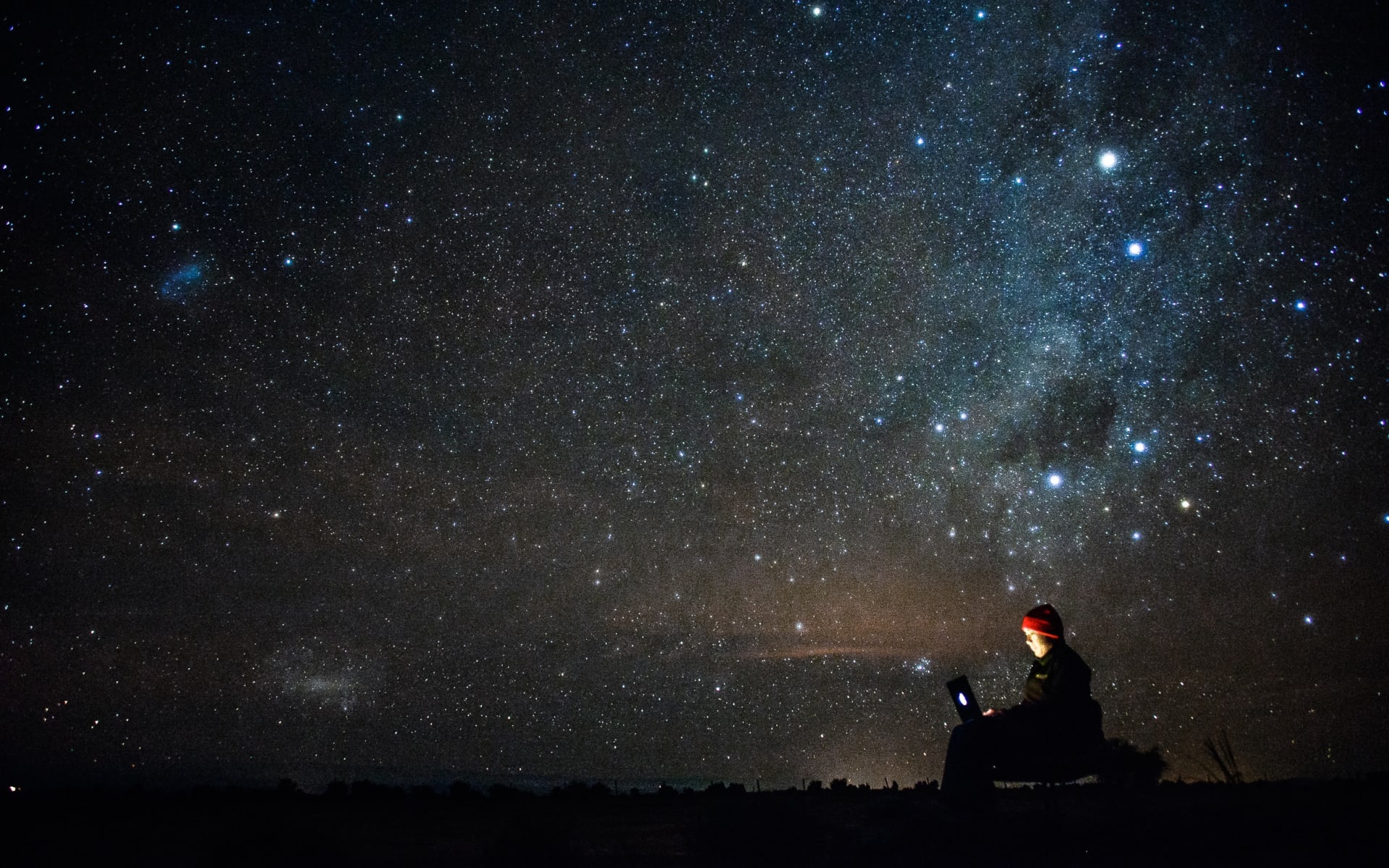A person sits with a torch under a black sky twinkling with thousands of stars. 
