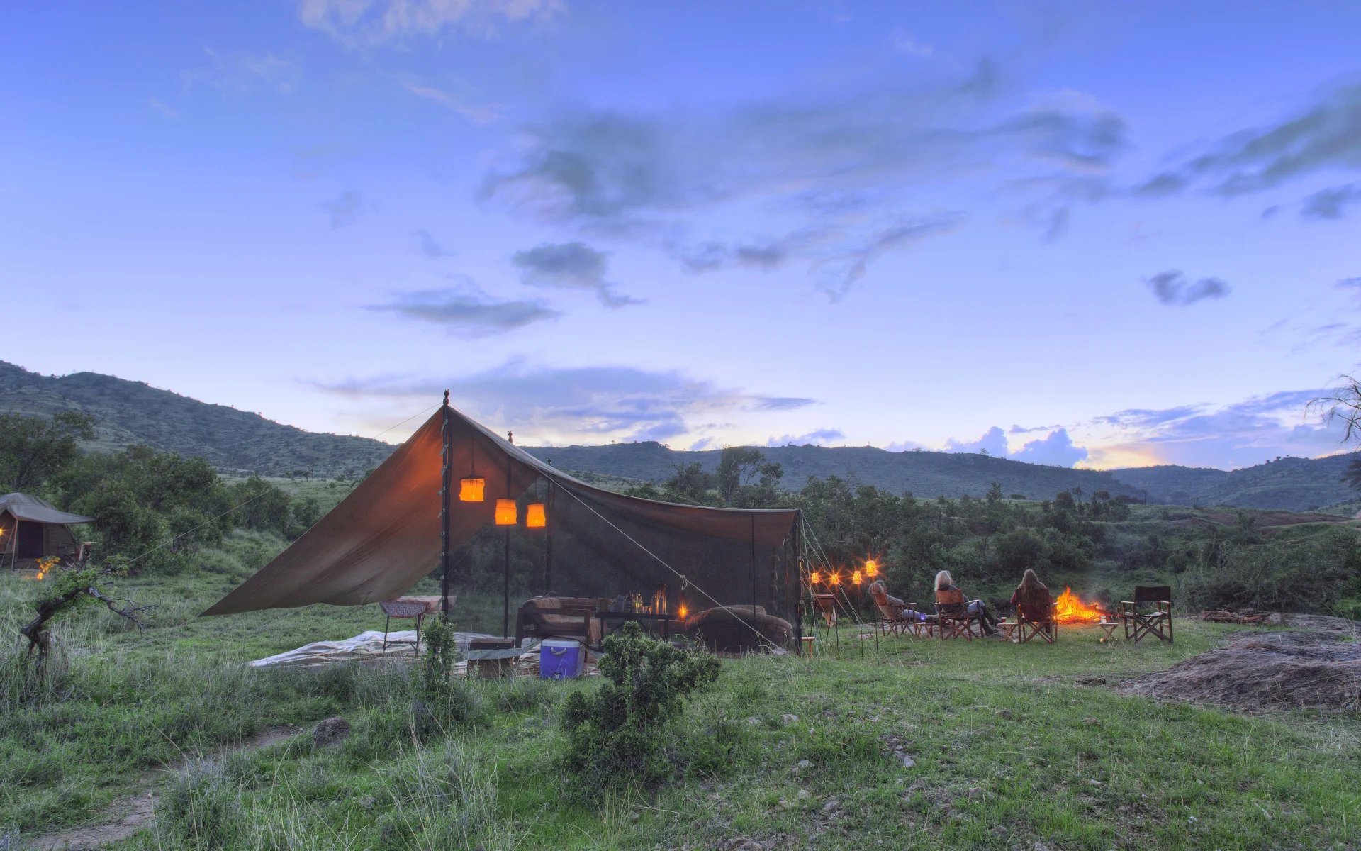 The image shows people fly camping during the early evening outside Borana Lodge. The tent is dimly lit by lamps and the scenery is mountainous.