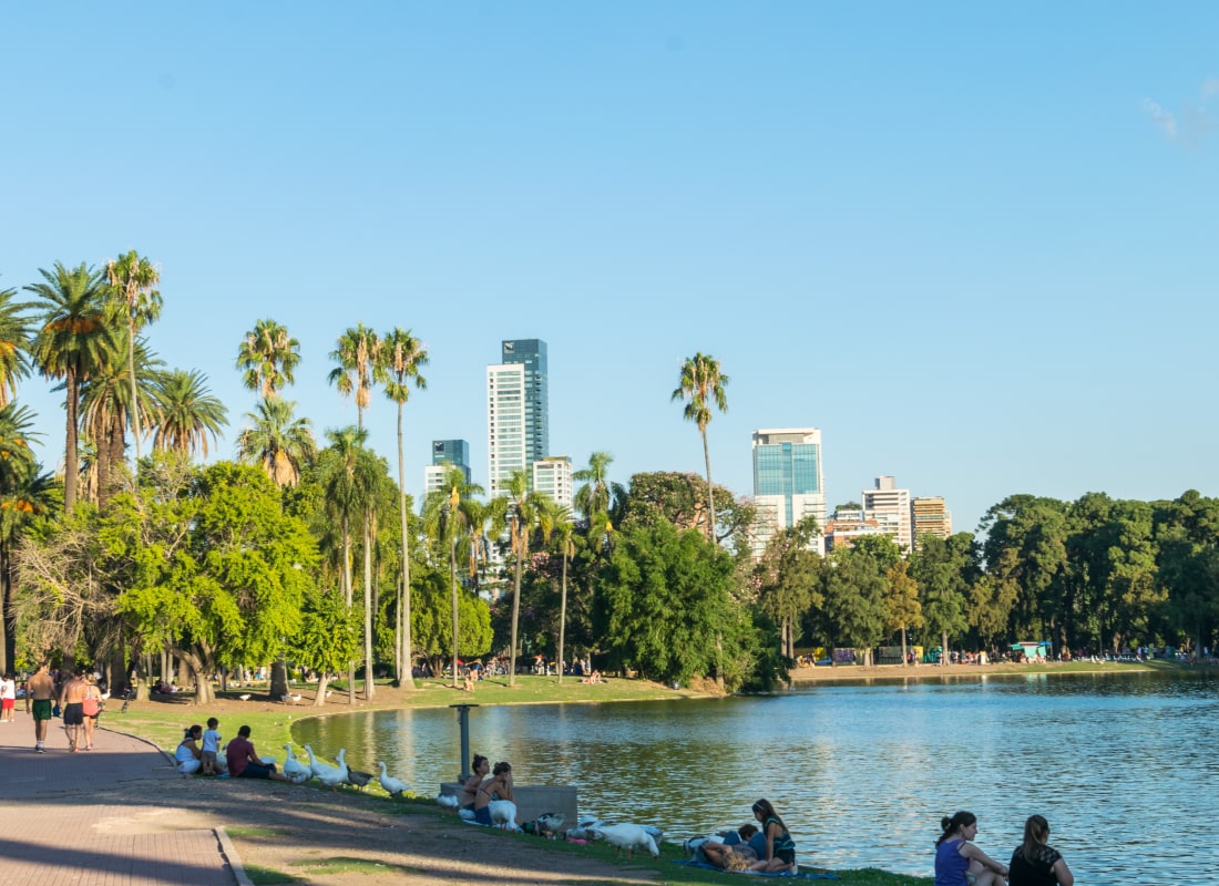 People are sitting in front of a lake with trees surrounding it and skyscrapers in the distance. 