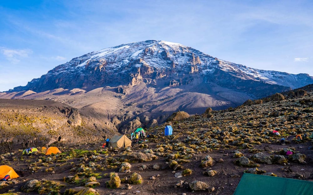 A tented camp is set up on a flat area near a snowy peak at Mount Kilimanjaro.