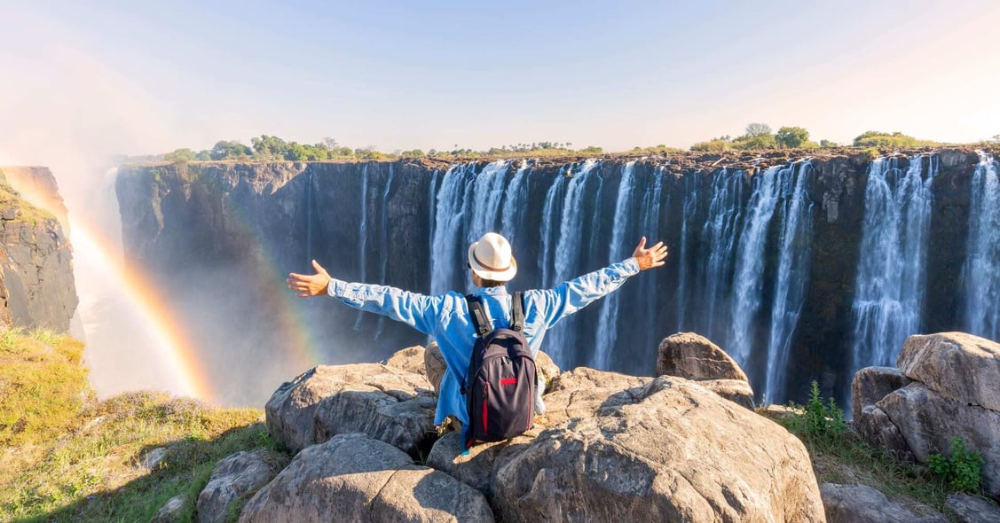 A traveller perches on a rock facing Victoria Falls during midday.