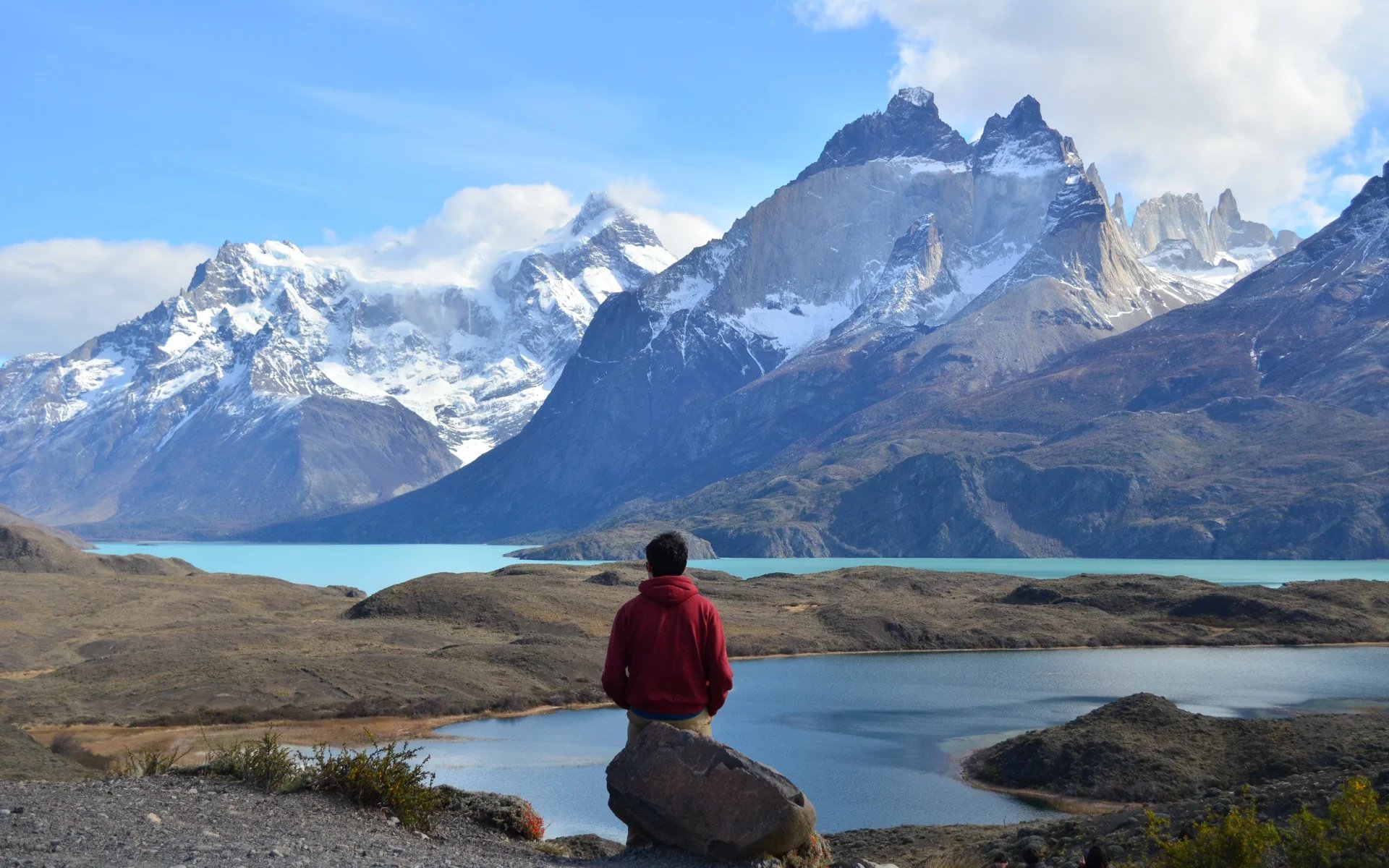 A man is standing in front of a lake and snow-capped mountains. 