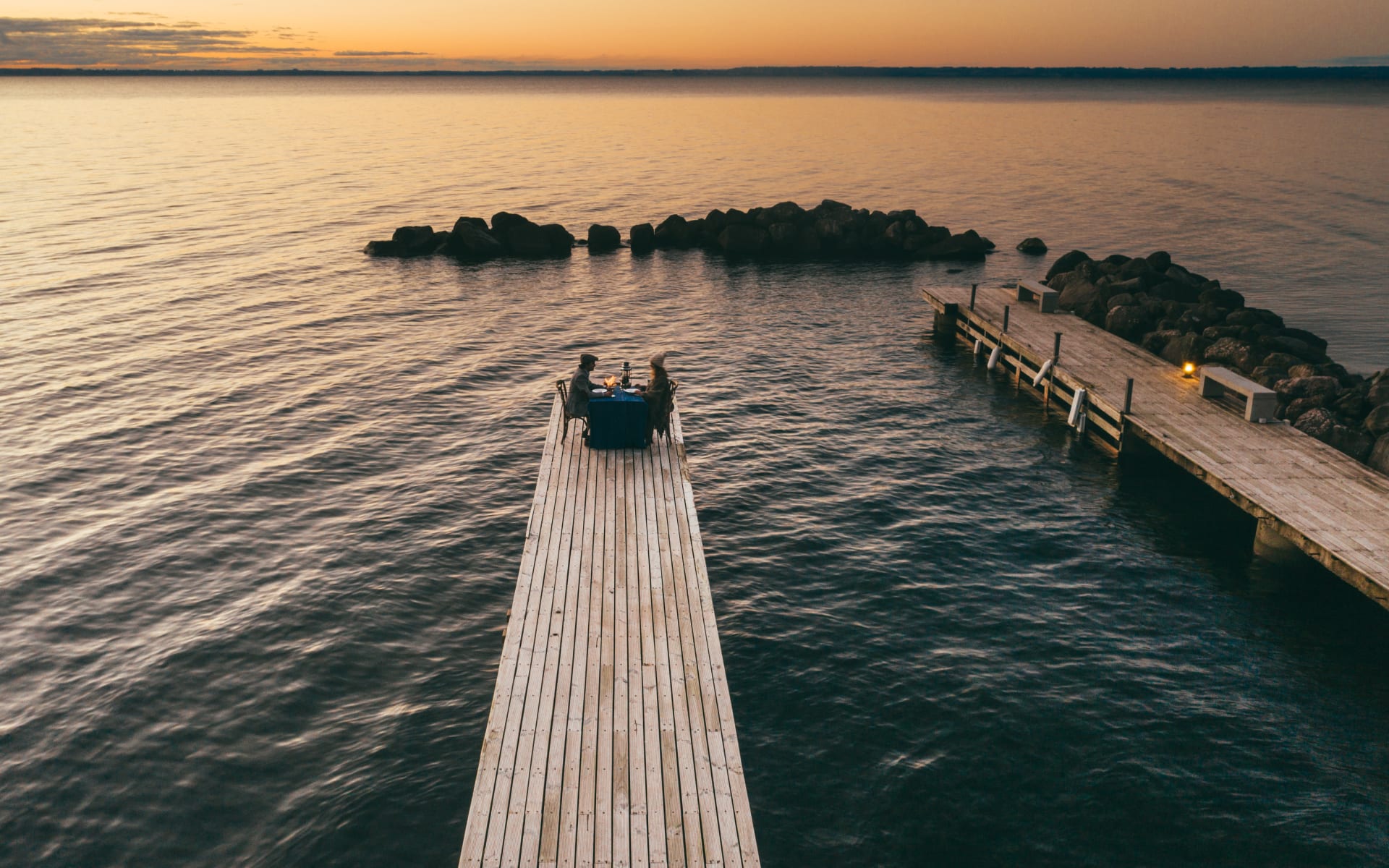 On the pier is a couple sitting at a table while the sun starts to set. 