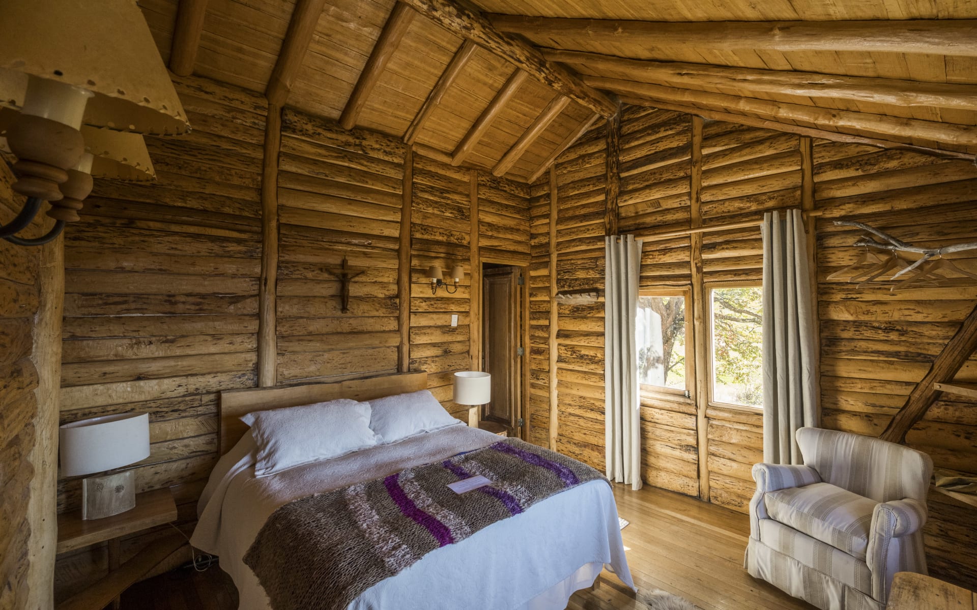 A double bed sits in the middle of a room comprised of wood, overlooking windows and an armchair. 