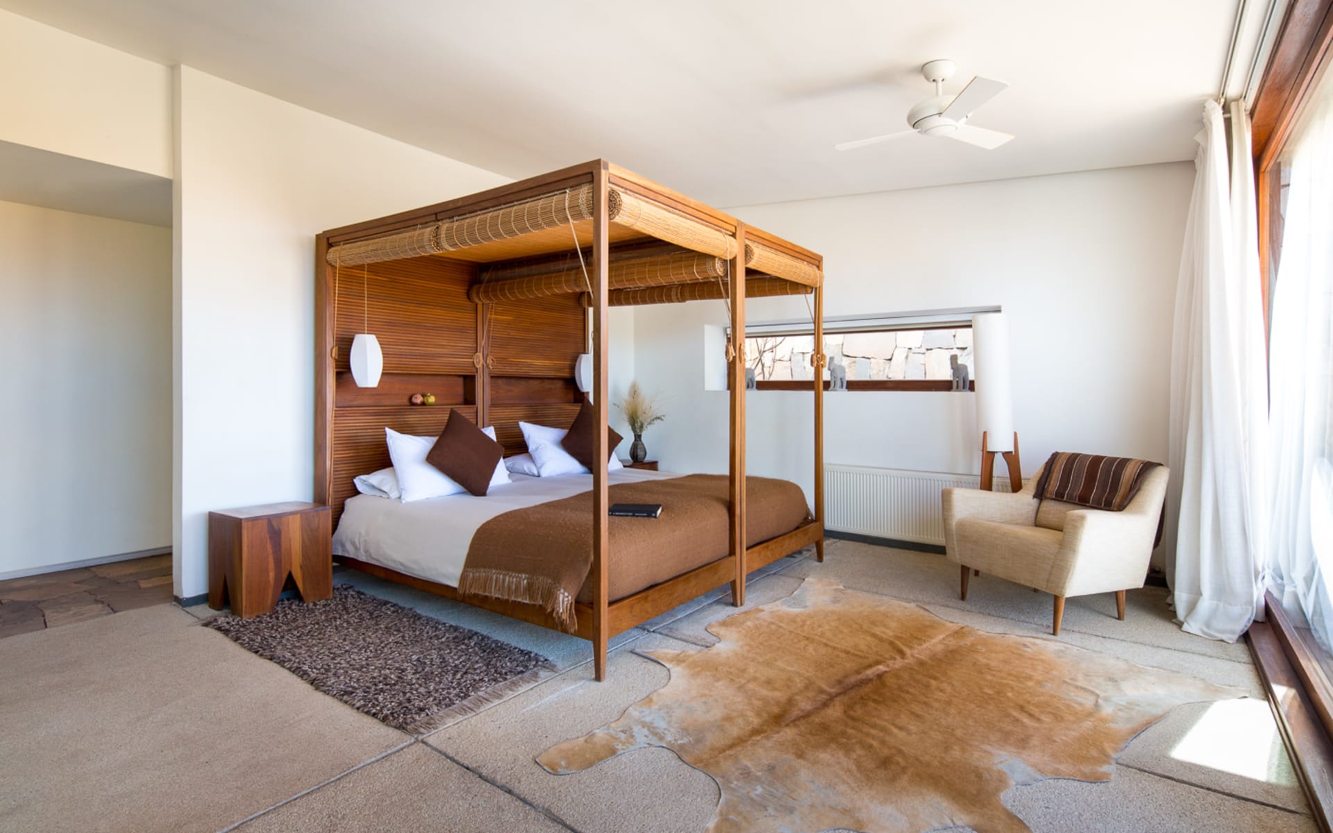 A canopy bed overlooks an earthy rug, white armchair and airy curtains. 