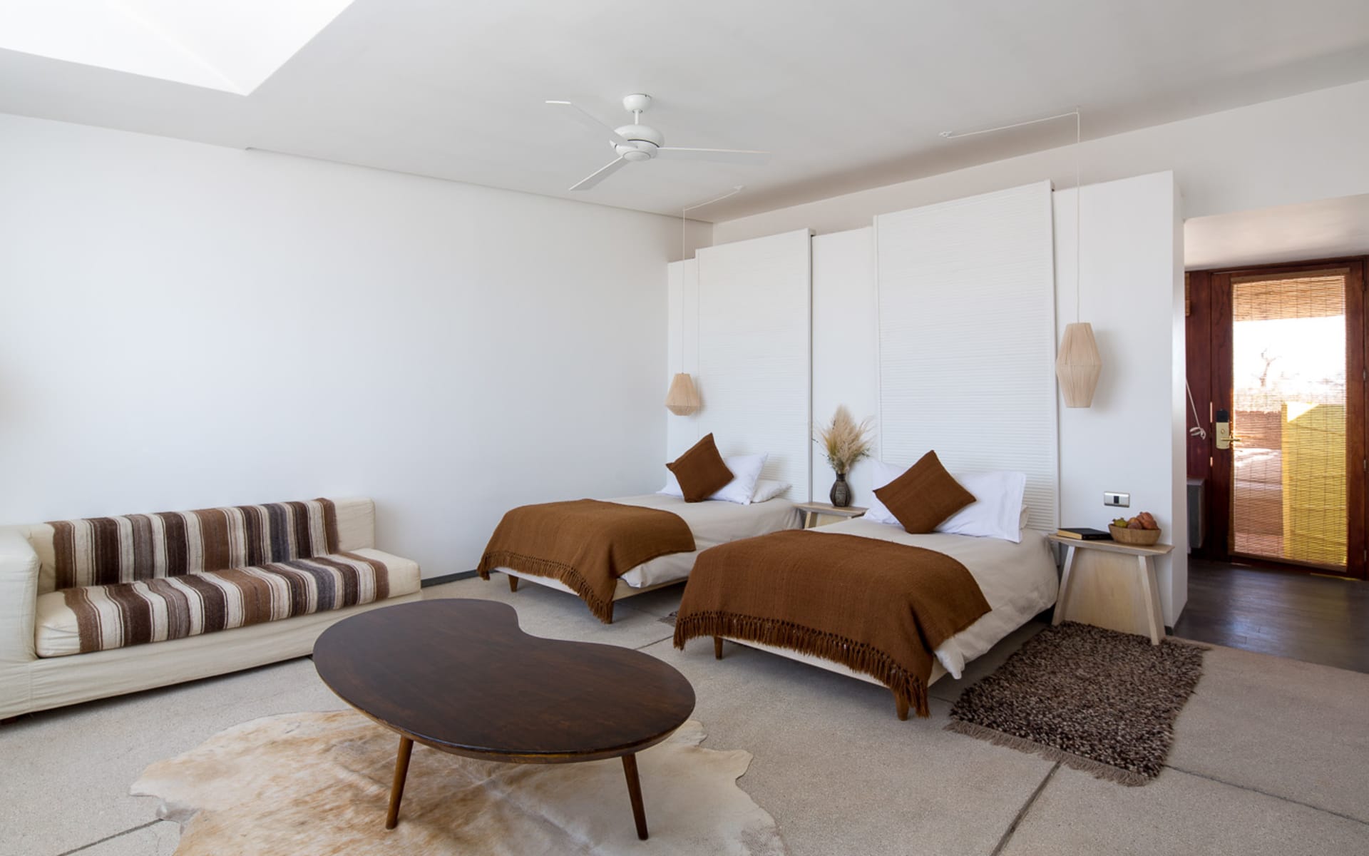 Twin beds have brown pillows and throw blankets and overlook a wooden coffee table and low sofa. 